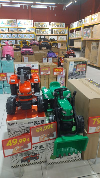 In a retail store in Pylaia Municipal Unit, Greece, a display of children's ride-on toys is presented. The foreground features two pedal-powered tractors, one orange and one green, positioned on top of their respective product boxes. The orange tractor is on the left, and the green tractor is on the right. Red signage with yellow lettering indicating prices of "49,99 €" and "69,99 €" is visible in front of the tractors. Behind these, a variety of other ride-on vehicles, including pink scooters and cars, as well as larger toy trucks and ATVs, are displayed on yellow shelving units that extend towards the back of the store. The environment appears to be a well-lit toy section within a larger retail establishment, with numerous cardboard boxes stacked on shelves.