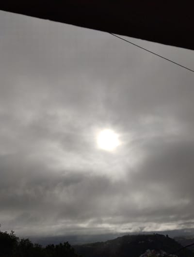 The image depicts an overcast sky with the sun partially visible through the clouds. The sun appears as a bright, diffused light source in the upper center of the frame. Below the sky, dark silhouettes of trees and hills are visible, suggesting a natural landscape. A dark, angled structure, possibly a roof or overhang, occupies the top portion of the image, casting a shadow. The overall impression is a moody, atmospheric scene, potentially depicting weather conditions common in Arucas, Spain, where such cloud cover and hilly terrain are found. There are no people or distinct objects identifiable in the scene beyond the natural elements and the man-made structure at the top.