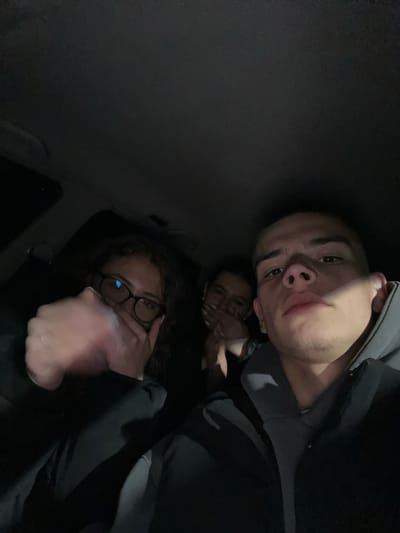 Three individuals are visible in a low-light environment, possibly the interior of a vehicle. The person in the foreground, a male with short hair, is positioned closest to the camera. Behind him, a female with glasses covers her mouth with her hand. In the background, a third person is partially obscured. The image does not provide enough information to determine its location in Karavelovo, Bulgaria.
