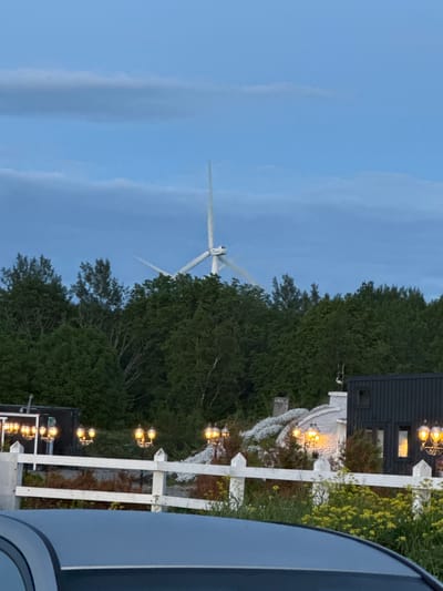 The image captures a twilight scene with a wind turbine visible above a dense line of trees. In the foreground, a white picket fence runs horizontally across the frame, separating the viewer from a low-lying building with multiple lit lamps. The building appears to have a dark, modern section and a lighter, possibly older, section covered in white flowers. A small portion of a vehicle's roof is visible at the bottom of the frame. The sky is a dusky blue with scattered clouds. The wind turbine, a prominent feature, suggests the location is likely in an area with renewable energy infrastructure. Based on its presence and the overall landscape, this scene is consistent with the outskirts of Paldiski, Estonia, a city known for its wind farms and coastal features.