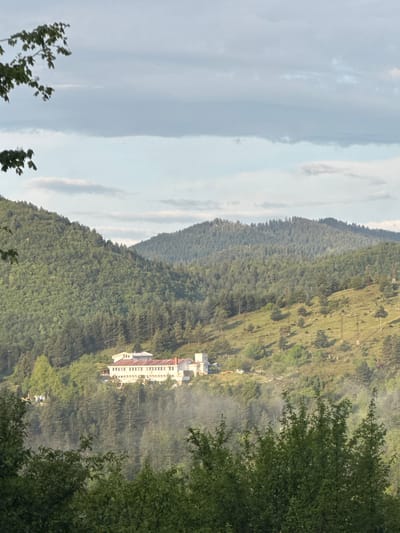 The image shows a landscape view in Strazha, Bulgaria. A white building with a red roof sits on a hillside among dense trees. The background features layers of green hills extending into the distance, with a strip of cloudy sky visible on the left. In the foreground, tree branches frame the right and bottom left of the image. The scene has a hazy, somewhat muted quality.