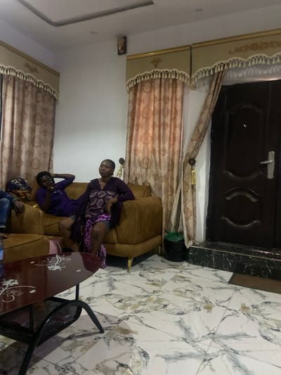 The image displays an interior scene in Ungongo, Nigeria, appearing to be a living room or lounge. Three individuals are visible. On a brown upholstered sofa, an individual in a purple top and dark bottoms is reclining, holding a dark object, possibly a mobile phone, with their right hand near their head. To their right, another individual, wearing a purple long-sleeved top and a patterned purple and white fabric draped over their lap, sits upright on the same sofa, looking slightly upwards and to the right. A third person is partially visible on the far left, wearing blue jeans. In the foreground, a dark-tinted glass-top coffee table with white decorative patterns and a black metal base is present. The room features white walls, a white ceiling with a recessed rectangular design, and white and grey patterned floor tiles. Ornate beige and gold curtains with decorative pelmets and tassels are drawn on the right wall and behind the sofa on the left. A dark brown door with silver hardware is visible on the right, next to which a black waste bin with a green lid is positioned. A wall sconce is mounted on the upper left wall.