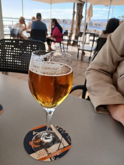 A vibrant outdoor scene captures a glass of golden beer, topped with a frothy head and covered in condensation droplets, resting on a smooth grey table. The glass is a stemmed tulip or snifter style, indicating a specific type of brew. Beneath its stem, a circular coaster with an abstract pattern in blue, orange, and white is visible, featuring an indecipherable logo or text.

In the immediate foreground, the arm and torso of a person wearing a light beige or cream-colored jacket are partially visible, suggesting they are seated at the same table as the beer. The background is softly blurred, showing a lively outdoor terrace setting. Several indistinct figures of people are visible, some seated at other tables, enjoying the sunny weather. A person in a bright pink top is noticeable among them. Black metal chairs, some with perforated designs, are scattered across the terrace.

Overhead, light-colored awnings or umbrellas provide shade, indicating bright daylight conditions. Beyond the terrace, a wide expanse of what appears to be a body of water, possibly the sea or a large harbor, stretches towards the horizon, framed by distant landmasses or buildings. The overall ambiance suggests a relaxed and leisurely moment, likely at a bar, restaurant, or cafe with a scenic view, consistent with a location like Agüimes, Spain, which offers coastal vistas.