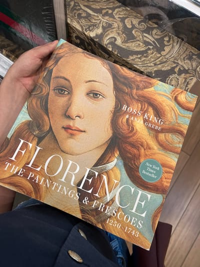 A person's left hand holds an open copy of the hardcover book "FLORENCE THE PAINTINGS & FRESCOES 1250 - 1743" by Ross King & Anja Grebe. The book's cover features a close-up of a female face with flowing reddish-blonde hair, reminiscent of Botticelli's "The Birth of Venus," and includes a teal "New York Times Bestseller" sticker. The person is partially visible, wearing a dark blue garment with two prominent silver-colored buttons over what appears to be blue denim pants. In the background, to the upper left, a book or box with a distinctive green, red, and green striped pattern, characteristic of Gucci branding, is visible, partially wrapped in plastic. To the upper right, another book or box displays a dark brown background with an ornate light brown/gold damask pattern. A dark red upholstered surface is partially visible below the book. The scene is located within a Dillard's department store in El Paso, United States.