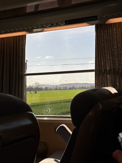 The image depicts a static interior view from a passenger vehicle, likely a train, looking outwards through a large rectangular window at a rural landscape in the vicinity of Pexiora, France. In the immediate foreground, dark upholstered seatbacks are visible on the left and right, partially obscuring the interior. The window is framed by dark brown, vertically textured curtains. Above the window, an overhead panel containing a series of circular vents or lights is visible, with segments marked "78" and "77." Bright sunlight enters from the exterior, casting distinct shadows from the seat structures and window frame within the vehicle.
The exterior scene reveals a vast, flat expanse of vibrant green agricultural fields under a clear, bright blue sky dotted with scattered, wispy white cumulus clouds. Several bare deciduous trees, likely in a dormant state, are sparsely distributed across the green field. Multiple thin, dark horizontal lines, consistent with utility wires or fences, traverse the upper portion of the field and lower sky. In the distant background, a low range of tree-covered hills or mountains stretches across the horizon. A circular reflection or smudge is present on the windowpane in the center-lower portion, overlying the green field.