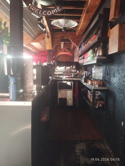 The image shows the interior of a bar or cafe. The perspective is from behind a counter looking down a narrow aisle. The bar has a wooden ceiling with exposed beams and a metal fan mounted on it. There are shelves on the right side stocked with bottles and cans, a large water jug, and various containers. On the left, there is a blue thermos, a menu, and some stainless steel equipment. A red cloth hangs on a piece of equipment. The floor is made of wooden planks. The scene is dimly lit with artificial lights. The date and time "19.06.2026 06:15" are visible in the lower right corner. There are no people visible in the image. The location is identified as Kiparisnoe, Ukraine.