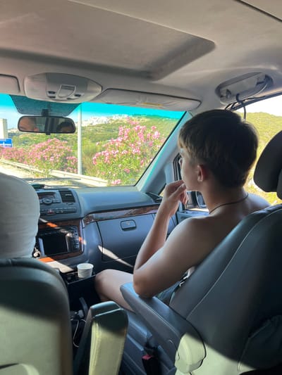 The image shows the interior of a car, with a young person in the passenger seat looking out the window. The person has short brown hair and is wearing a black necklace. The car's dashboard and a cup holder with a patterned cup are visible. Outside the car, a landscape with pink flowers and buildings on a hill can be seen, suggesting the location is Nea Peramos, Greece. The light suggests it is daytime. Another person is partially visible in the driver's seat, reflected in the rearview mirror.