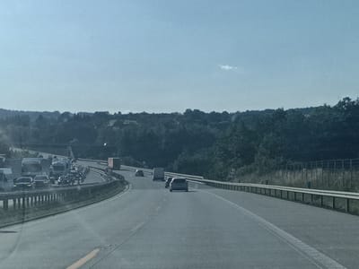 The image captures a highway scene in Hrusice, Czechia, likely during the daytime given the bright sky and overall illumination. The weather appears clear with a few wispy clouds visible against a pale blue sky. The road is a multi-lane highway, with a noticeable traffic jam ahead. Several vehicles, including cars and trucks, are stopped or moving very slowly. The road is lined with metal guardrails and vegetation. In the background, a dense forest covers rolling hills. There is a bridge or overpass visible in the distance on the left.
Notable details include traffic signs along the roadside, one of which appears to be a circular sign with a symbol inside, possibly indicating a speed limit or restriction. The overall impression is one of congestion on a typical highway day, surrounded by natural landscape. No people are clearly discernible in the image, but the presence of numerous vehicles implies human activity. The setting is a rural or semi-rural area, as evidenced by the surrounding greenery and wooded hills.