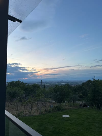 The image depicts a landscape scene, seemingly taken from an elevated position, potentially a balcony or high window, in the city of Brestnik, Bulgaria. The foreground features the corner of a glass panel, possibly part of a balcony railing, with water droplets indicating recent rain or condensation. The background reveals a panoramic view. The sky is a mix of blue with scattered clouds at various altitudes. There are hints of sunset hues, with orange and pink light near the horizon. Below the sky, a cityscape stretches into the distance, suggesting a populated area. The terrain appears hilly, with trees and vegetation visible. A small, circular white object is seen on a grassy lawn. There are no people are clearly visible.