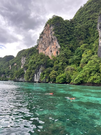 Two individuals are snorkeling in the clear, turquoise waters off the coast of Ko, Thailand. They are both wearing orange snorkeling vests and masks, indicating they are actively engaged in the activity. The water is remarkably transparent, revealing the rocky seabed below. Towering, verdant cliffs rise dramatically from the water's edge, covered in lush green vegetation. The sky above is overcast with dramatic gray clouds, suggesting a day with variable weather conditions. In the distance, a small boat is visible near the shoreline, adding a subtle human presence to the vast natural landscape. The overall scene captures a serene moment of recreational activity within a breathtaking natural environment.