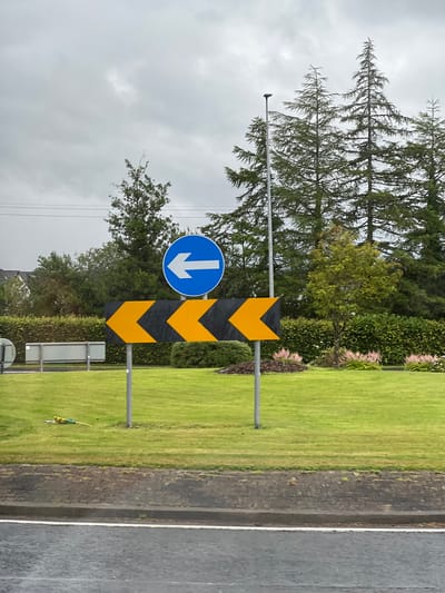 The image shows a street sign with a downward-pointing blue arrow and a chevron pattern of black and yellow stripes, likely indicating a turn or a hazard on the road. The sign is mounted on a metal pole in what appears to be a roadside setting. To the left of the sign, there are trees, including tall coniferous trees and a deciduous tree with green leaves. A well-maintained green lawn is visible to the right of the sign, bordering a paved surface that could be a road or a pavement. The sky is overcast, suggesting a cloudy or rainy day. The overall scene is a typical roadside environment, possibly in a rural or suburban area. There is no visible text in the image, other than the symbols on the sign itself. The image is captured in a vertical orientation.