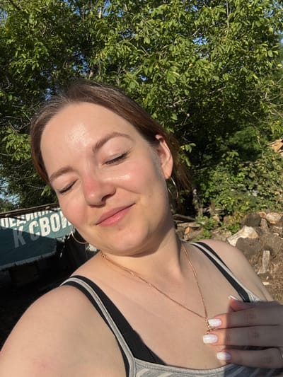 A woman is pictured in an outdoor setting, likely in a rural area given the trees and woodpile in the background. The woman has her eyes closed and is smiling, suggesting a moment of enjoyment. She is wearing a grey and black tank top, a rose gold necklace with a pendant, and hoop earrings. Her nails are painted white and appear to have a glitter finish. In the background, there is a green tarp with white lettering that reads "К СВОИМ". The image appears to be a selfie, taken in sunlight. The location in the city of Zubov, Russia, is not explicitly indicated by any visual cues in the image.