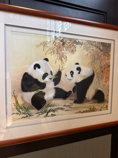 This is a framed artwork featuring two pandas in a naturalistic setting. The panda on the left is sitting and appears to be eating bamboo shoots. The panda on the right is also sitting, looking towards the other panda. The background depicts a stylized representation of trees with reddish-brown leaves and some green foliage. The artwork is framed with a wooden border. The image appears to be taken indoors, with reflections visible on the glass of the frame. The location is not determinable from the image itself.