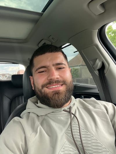 A man with a beard and short dark hair is looking at the camera, smiling. He is wearing a light gray hooded sweatshirt. He is sitting in a car, with the sunroof open above him. The view through the sunroof shows trees and a cloudy sky. The interior of the car is visible, including the headrest of the seat in front of him. The image is taken from a low angle, looking up at the man and the sunroof. The image appears to be taken during the daytime. There is no visible text. The location context provided is Sânnicoară, Romania.