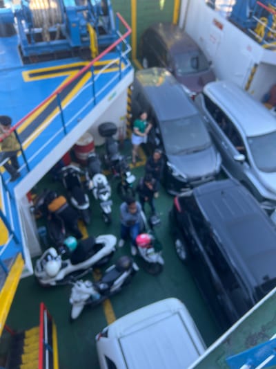 The image depicts a ferry filled with vehicles and people, likely in Ketapang, Indonesia. The scene is shot from above, looking down onto the deck of the ferry. Several cars are parked tightly together, including a white van on the right and a black SUV. Scattered among the cars are numerous motorcycles, some with riders sitting on them, others parked. A few people are visible, some standing or sitting near the vehicles, while others appear to be passengers.
The lighting suggests it could be daytime, but the overall image is quite blurry, making it difficult to discern specific details like the weather or precise time of day. The presence of many vehicles and people indicates a busy ferry crossing, a common mode of transport in Indonesia for connecting islands.
Notable details include the diverse mix of vehicles, from cars to motorcycles, suggesting a mix of travelers. Some individuals are wearing casual clothing, and one person is seen wearing a helmet, which is a common safety practice for motorcycle riders. The overall atmosphere appears to be functional and practical, typical of a ferry journey with a focus on transporting people and their vehicles. There is no discernible text in the image. The composition and the blurry nature of the image give it a candid, almost snapshot-like quality.