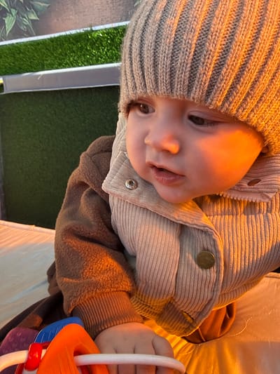 A baby wearing a knit hat and a corduroy jacket is lying on a white surface. The baby is holding a colorful toy. The background includes a patch of artificial turf. The location is unknown, and there is no verifiable information to indicate it is Кипчак, Russia.