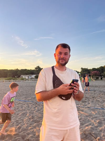 A man with a beard and short dark hair, wearing a white t-shirt, light-colored shorts, and a black fanny pack, stands on a sandy beach in Pistula, Montenegro. He is holding a smartphone in his hands and looking at the camera. To his left, a young boy in a purple t-shirt and dark shorts runs on the sand. In the background, several other people are visible, along with a playground structure and trees. The sky is a pale blue with some wispy clouds, and the setting sun casts a warm glow on the scene. There are also what appear to be portable toilet structures visible in the background, along with a wooden fence.