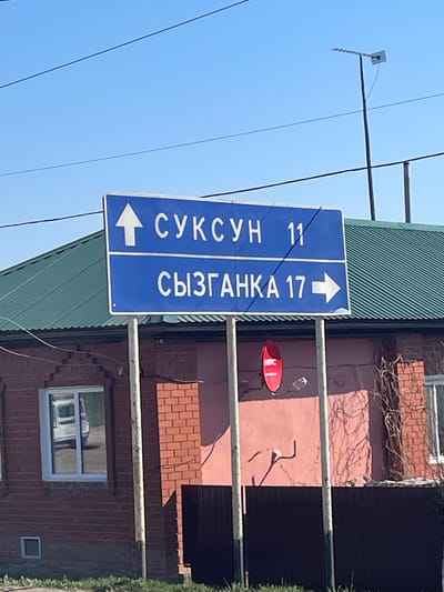 A medium shot shows a blue street sign in Поедуги, Russia. The sign is vertically oriented with white lettering indicating two destinations and distances. To the left with an arrow pointing left, it says "СУКСУН 11". To the right with an arrow pointing right, it says "СЫЗГАНКА 17". The sign is affixed to a metal pole.
Behind the sign, there is a building with a green corrugated metal roof and a brick facade. A red satellite dish is visible on the wall. A window is present on the brick wall of the building.
In the upper left of the frame, there are utility lines and an antenna against a clear blue sky. The image appears to be taken during the daytime.