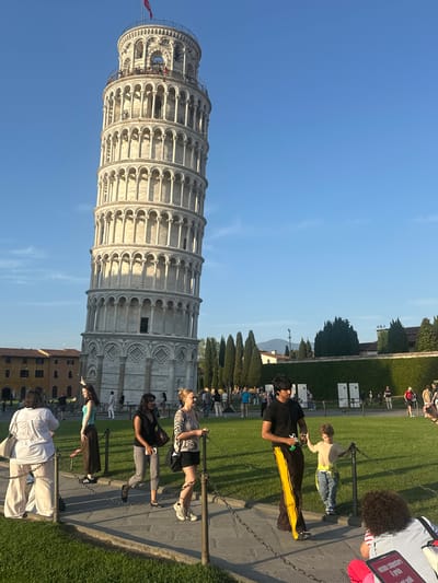 The image displays a bright, sunny scene at the historic site of the Leaning Tower of Pisa in Italy. The iconic white marble bell tower, featuring multiple levels of arched colonnades, dominates the left side of the frame, clearly exhibiting its famous lean. A small red flag is visible at the very top of the tower.

In the foreground and midground, numerous people are present, enjoying the tourist destination. A paved pathway runs horizontally across the scene, separated from a lush green lawn by a low chain barrier supported by metal posts. Several individuals are gathered along this barrier; in the immediate foreground, a man in a black shirt and black pants with a prominent yellow stripe stands holding a phone, while another person in a beige shirt and jeans leans against a post. Other visitors are seen walking, sitting on the grass, and observing the tower.

The clear blue sky and strong shadows indicate bright daylight, likely late afternoon or early evening. Beyond the main area, various trees and some distant buildings are visible. Several white informational signs are positioned on the grass, one of which partially displays the text "valuable culture." The overall atmosphere is that of a bustling tourist attraction.