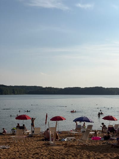 A wide shot captures a beach scene on a body of water, possibly a lake, with a treeline in the background. The sky is a clear, pale blue with faint wisps of clouds. The water is calm, with several people swimming or wading. On the sandy beach, various individuals are sunbathing, relaxing on lounge chairs, or walking. Red and blue umbrellas provide shade for some beachgoers. The overall impression is a typical summer day at a recreational area. This location is identified as being in the Ozyaritska-Slabadski rural council, Belarus.