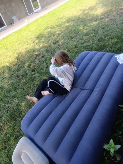 In Dumeni, Moldova, a young person with shoulder-length brown hair sits on an inflated navy blue mattress outdoors on a grassy lawn. The person is wearing a light-colored long-sleeved shirt and black pants with a white stripe down the side. Their left arm is raised to shield their face from the sun, and their bare feet are visible. A white cloth is on the upper right corner of the mattress. The background shows a gray building with windows and a paved walkway adjacent to the grass. Sunlight casts shadows on the grass.