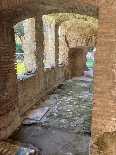 The image shows an interior view within Hadrian's Villa in Tivoli, Italy. The space is constructed of aged brick and stone, exhibiting significant decay. Natural light penetrates through openings in the structure, illuminating the interior. There is exposed masonry and visible signs of aging such as moss growth and crumbling material. A fragment of decorative tilework can be seen set within one of the wall sections, indicating the original presence of more elaborate design. Views to the exterior through the dilapidated structure reveal foliage and scaffolding. No people are visible.