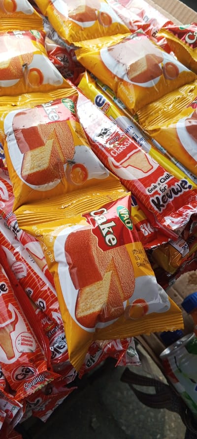 The image displays a close-up, elevated view of multiple individually packaged snack items, likely on a market stall or counter in Awjaw, Nigeria. The primary subjects are yellow and red foil-wrapped packages. The most prominent packages are yellow, labeled "WAO Cake," featuring an image of two slices of cake with a visible crumb structure and an egg yolk. Text on these packages includes "Net Content: 20g" and mentions a "thick milky flavor." A fly is visible resting on one of these yellow "WAO Cake" packages. Interspersed and beneath these are red and white striped packages, some identifiable as "Classic" brand, with text such as "Grab a Gala for Kewwa" and "Enriched with Vitamins & Minerals," depicting a sausage roll. The snacks are stacked in a disorganized pile. The background is blurred and dark, revealing a dark surface, a portion of a metallic can, and a blue plastic cap on an unseen bottle, indicating a commercial or vending environment. No individuals are visible.