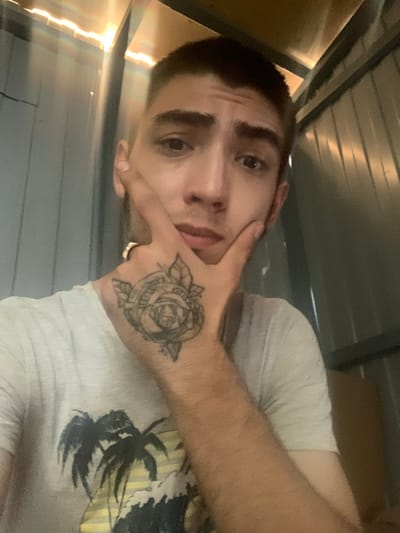 A close-up, over-the-shoulder shot shows a young person in a white t-shirt with a palm tree print. Their left hand is raised to their face, fingers touching their cheek, revealing a tattoo of a rose on the back of their hand and a gold ring on their index finger. The person's short, dark hair is visible, as are their dark eyebrows and eyes. The background appears to be an industrial or storage space with corrugated metal walls and overhead lighting. There is no information to indicate this image was taken in Kokhma, Russia.