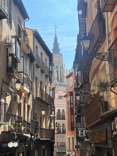 The image depicts a narrow, sunlit street in the city of Toledo, Spain. The street is framed by multi-story buildings on both sides, leading the eye towards the prominent, intricate Gothic bell tower of the Cathedral of Toledo in the background.
The buildings lining the street exhibit varied architectural styles and colors. On the left, facades are predominantly light beige, featuring multiple dark wrought-iron balconies, some enclosed glass balconies, and traditional black street lanterns mounted on ornate iron brackets. An external air conditioning unit is visible on one building's facade. Strong shadows are cast on this side, indicating sunlight from the upper right.
On the right, buildings display warmer tones including orange, cream, and brown. They also feature wrought-iron balconies, some adorned with potted plants, and additional decorative street lanterns. A prominent vertical sign for "HOSTAL H" is visible on a building on the right, along with smaller commercial signs. In the midground, between the immediate street buildings and the Cathedral, other structures with arched windows and varied facade colors are discernible.
The sky above is bright blue with scattered white clouds. No individuals or active subjects are visible within the frame.
