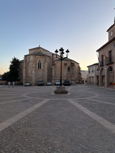 The image shows a town square in Salmerón, Spain. A large stone church, possibly a cathedral, dominates the left side of the frame. In front of the church, several cars are parked. A prominent street lamp stands in the center of the square. To the right of the church, a building with arches and balconies, likely a town hall, is visible. The ground of the square is paved with cobblestones, arranged in a patterned design. Two men are standing and conversing in the foreground on the left. The sky is clear and bright, indicating daytime.