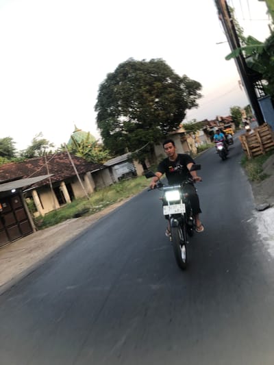 In Glagah Arum, Indonesia, a man is riding a black motorcycle down a paved road. He is wearing a black t-shirt and shorts, with white sandals. The motorcycle has a white license plate. In the background, there are houses with red tile roofs, a large tree, and a green domed building, possibly a mosque. Another person is riding a motorcycle further down the road, and another person is visible in the distance. A utility pole with wires stands on the right side of the road, next to stacked wooden pallets. The sky is overcast.