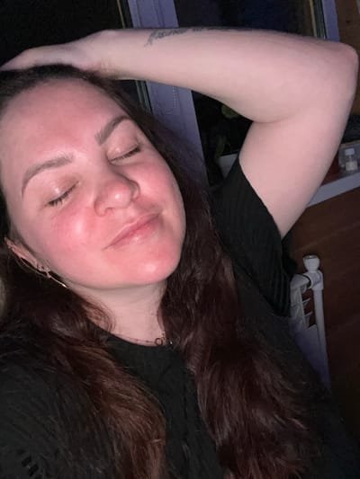 A woman is lying down, eyes closed. Her head is tilted to the side, and one arm is raised above her head. She has dark hair and is wearing a black top. The background includes a radiator and some bedding. The location is not identifiable in the image, so Bol'shakino, Russia, cannot be confirmed.