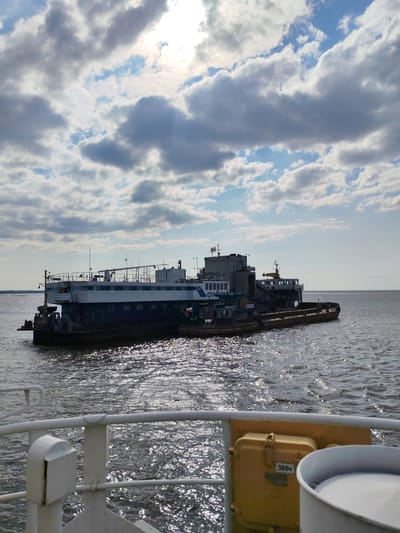 The image captures a vessel docked on a body of water, likely the Sheksna River, near the former village of Krokhino in the Vologda Oblast of Russia. The vessel appears to be a large barge or a specialized workboat, with a multi-level superstructure. The barge has a dark hull and a lighter upper structure with multiple windows. There is a flag on the upper deck. The water is choppy, with sunlight glinting off the surface. The sky is partly cloudy, with patches of blue sky visible. In the foreground, the railing and part of another vessel are visible, suggesting the photo was taken from another boat. The overall scene is one of industrial activity on the water, with the vastness of the river and sky dominating the background.