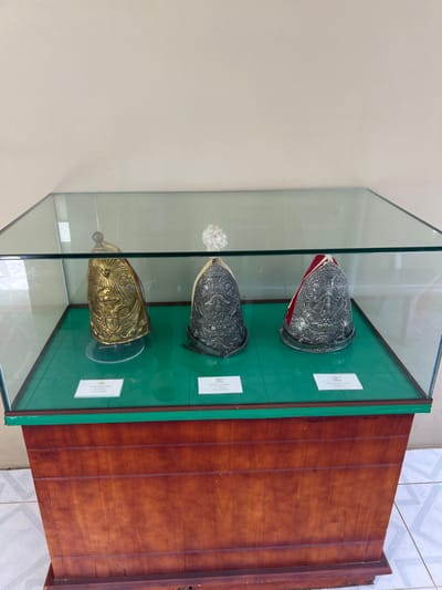 A glass display case houses three ornamental hats or helmets, each on a green platform with an accompanying label. The top two helmets are silver, adorned with intricate designs, one featuring a red trim. The bottom helmet is gold in color and design. The display case sits atop a brown wooden cabinet with vertical paneling. The background includes a light cream wall and a section of tiled flooring.