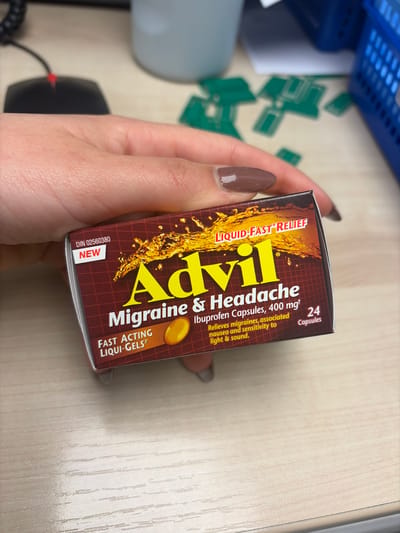 A hand is holding a box of Advil Migraine & Headache medication. The box is brown with yellow lettering and images of liquid-filled capsules. Text on the box includes: "LIQUID FAST RELIEF," "Advil Migraine & Headache," "FAST ACTING LIQUI-GELS," "Ibuprofen Capsules, 400 mgt," and "24 Capsules." The hand has polished nails. The background includes a desk with various objects, including a blue basket, green objects with geometrical shapes, a computer mouse, and a light-colored desktop surface. It is impossible to determine if this image was taken in the city of Longueuil, Canada, as there is no information to infer this.