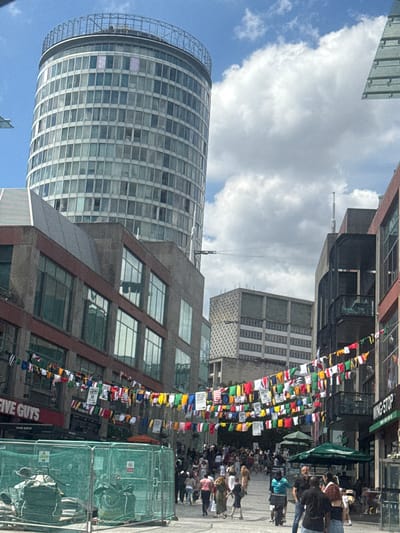 The image depicts a street scene in Birmingham, United Kingdom, likely near the Rotunda building, a prominent circular glass tower visible in the background. The street is lined with multi-story buildings, some featuring brick and concrete facades, while others are modern glass structures. A vibrant canopy of colorful flags, possibly representing various nations or sporting events, stretches across the street, creating a festive atmosphere.
Below the flags, storefronts are visible, including a "Five Guys" restaurant. The street is bustling with people, with individuals and small groups walking, some dressed casually. A few people are seen interacting, and one person is on a scooter. In the foreground, a construction site is cordoned off with green safety fencing, revealing construction materials and equipment. The sky is partly cloudy, with a bright blue sky peeking through. The overall impression is of a lively urban environment on a bright, possibly summer day.