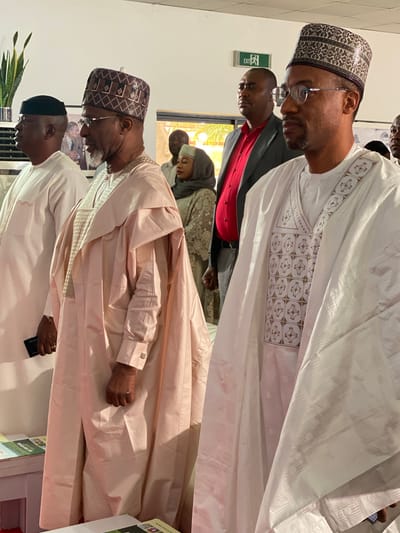 The image depicts a group of people indoors, likely in Kwarau, Nigeria. Several men are prominently featured, dressed in traditional Northern Nigerian attire, including elaborately embroidered gowns and caps. One man wears a white gown with detailed geometric patterns, while another sports a light pink gown. Some of the men wear glasses and are looking down. A woman in a head covering is partially visible in the background. An emergency exit sign is visible on the wall. The environment appears to be well-lit, possibly a meeting or gathering space. A man is seen holding what appears to be a cellphone. A stack of papers or leaflets are visible.