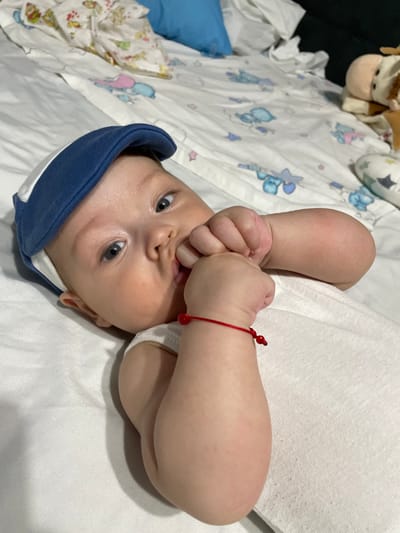 The image shows a close-up, overhead view of a baby lying on a white surface, possibly a bed or changing table, adorned with a blue and white patterned sheet featuring cartoon animals like bears and stars. The baby is wearing a white onesie and a blue and white soft hat, tilted slightly to the side. Their hands are brought up to their mouth, with fingers in their mouth, suggesting they might be teething or exploring their hands. The baby has light skin, bright blue eyes, and light brown hair. A thin red string bracelet with a small red bead is visible on their right wrist. In the background, partially out of focus, are some stuffed animals and a blue pillow. The lighting suggests an indoor setting, likely during the day, with no discernible weather conditions. The overall mood is tender and intimate, capturing a moment of infancy. There is no visible text in the image. The location context is Fălești, Moldova.