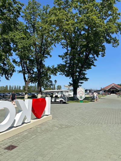 In the city of Koson, Ukraine, a sunny day presents a paved promenade with large white lettering spelling "I LOVE KOSON," the word "LOVE" represented by a red heart. A white golf cart is parked nearby, and several people are seen strolling or standing in the background. Beyond the promenade, a parking lot is visible with cars, and large trees with green foliage cast shadows. Further in the distance, a building with a red roof and a sign that reads "СТОРІНКА" is situated. The sky is clear and blue.