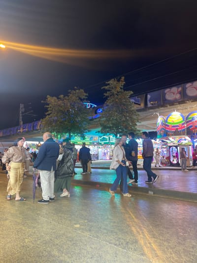 At an amusement park in Jerez De La Frontera, Spain, people are milling about in the early evening. Individuals are strolling, some in groups, on a paved surface. Trees are interspersed among the amusement park attractions. The night sky is visible. A beam of light shoots upward.