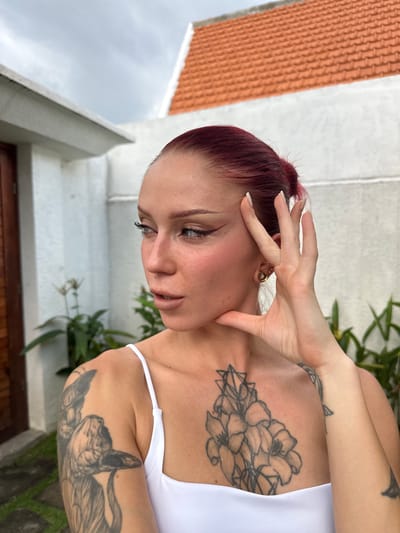 An outdoor scene in Kerobokan, Indonesia, features a female individual shown from the chest up, positioned slightly to her left, with her gaze directed towards her left (viewer's right). She has reddish-brown hair, pulled back, and light-colored eyes with prominent black winged eyeliner and mascara. Her cheeks exhibit blush, and her lips are tinted light brown. Her right hand is raised, with fingers splayed, the thumb resting along her jawline, and the index and middle fingers touching her temple. Her fingernails are long and painted white. The left earlobe displays a gold-toned ear tunnel, containing a smaller gold stud. She wears a white top with thin straps. Visible tattoos include two sphinx cats on her left upper arm, a large floral and geometric pattern on her upper chest, and a faded design resembling a marine creature's tail on her inner left forearm.
The background consists of a light-colored, textured wall. To the immediate left, a brown wooden door or gate is partially visible. Green tropical foliage is present behind and to the sides of the individual. Above the wall in the upper right quadrant of the image, a section of an orange-tiled roof is visible. The sky overhead is overcast and grey. A paved ground surface with stone-like elements is partially visible in the lower left.