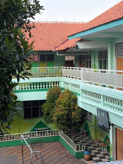 The image captures the exterior of a light green school building in Pekalongan, Indonesia, under an overcast sky. The building features multiple levels with balconies and walkways, all adorned with white railings. The roof is covered in a classic red tile pattern. On the ground floor, a checkerboard tiled area is visible, with a blue and brown barrel, a small bench, and a basketball hoop mounted on the wall. The courtyard pavement is a mix of red and yellow interlocking bricks. Lush green trees and foliage are present, with one particularly prominent tree in the center of the frame. The walls of the building are painted in varying shades of green and feature a mural-like design. There are no people visible in the image.