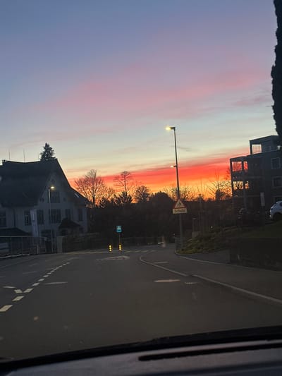 The image depicts a street scene in Wetzikon (ZH), Switzerland, at dusk. The sky is painted with pink and orange hues, indicating either sunrise or sunset. On the right side of the road, there is a two-story building, and further ahead, a "Schule" (school) sign is visible. A building with multiple windows is across the street. Streetlights are illuminated, casting a glow on the road surface, and white markings define lanes on the road. The photo seems to be taken from inside a vehicle, as part of the dashboard is visible on the right side.