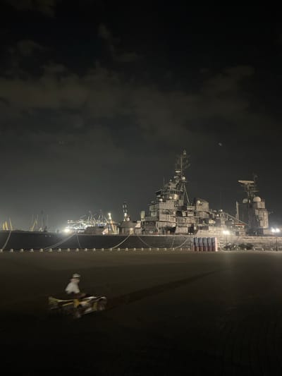 This is a vertically oriented, nighttime photograph taken from a slightly elevated position, likely a dock or pier. The primary subject is a large, complex ship, possibly a warship or a large freighter, docked at night. The ship is heavily lit with numerous deck lights, illuminating its many decks, superstructure, radar arrays, and antennas. The ship's hull is dark and appears to be made of metal. Some cargo, possibly containers, are stacked on the deck.
To the right of the ship, there is a dark, flat surface, likely the rest of the pier or dock. On this surface, a small object that appears to be a person on a motorized scooter or similar vehicle is visible in the lower right portion of the frame. The person and vehicle are illuminated by the ambient light, and the person appears to be wearing light-colored clothing.
The background is dark, with some lighter areas suggesting the night sky and possibly clouds. The overall scene conveys a sense of activity at a port or naval base during the night. The lighting on the ship creates a dramatic contrast with the dark surroundings. The presence of the ship, the cargo, and the dock infrastructure suggest a functional, industrial setting. The activity of the person on the scooter indicates some level of ongoing operation or movement on the dock. The time of day is clearly night. The weather appears to be clear enough for visibility, though some clouds might be present.
There is some visible text on the stacked cargo on the ship, appearing as red and blue blocks with white lettering. It's difficult to discern the exact words, but the presence of "РОССИЯ" (Russia) is partially visible.