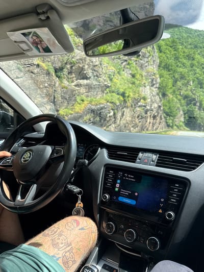 The image provides an interior view from a car, showcasing the dashboard and a driver's leg, set against a backdrop of a lush, rocky mountain landscape. The scene suggests a road trip or scenic drive through mountainous terrain, consistent with the provided location context of Dorkovo, Bulgaria.
The car's interior features a modern dashboard with a steering wheel bearing a Skoda logo. The infotainment system is prominent, displaying a navigation map with text in Cyrillic. The screen shows the time "14:45" and "4G" network status. It indicates a destination or point of interest "+15 km" away on "ул. Хан Аспарух" (ul. Khan Asparuh), with a message stating "Скоростта намалява" (Speed decreases) "След 180 м" (After 180 m) and a "Затвори" (Close) button. Below the screen are physical buttons for functions such as "NAV", "APP", "CAR", "MENU", "RADIO", "MEDIA", "PHONE", "VOICE", "AUTO", "A/C", "SYNC", "OFF", and "Air Care". Air vents and a hazard light button are also visible.
To the left, a rearview mirror is seen, and a sun visor has a small photograph of two smiling people taped to it. The windshield frames the exterior view of a steep, green-covered rocky hillside under what appears to be a bright, sunny day.
A person's leg, heavily adorned with various cartoon-like tattoos, is visible in the foreground near the gear shifter, which is marked "DSG". A set of car keys with a remote fob and a black keyring with a white graphic hangs nearby. The digital instrument cluster displays "14:44" and a speedometer with markings up to 220 km/h, showing "185" which likely refers to remaining range. The "PASSENGER AIRBAG OFF" indicator is also lit.