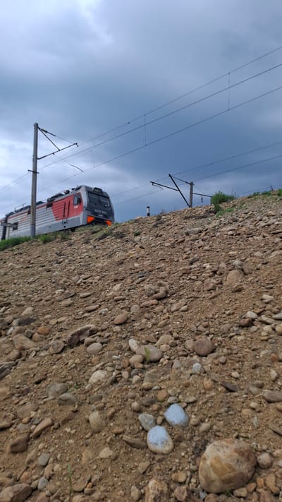 An overhead shot captures a Russian train, specifically a 33CSK 1600 model, traveling on a rocky embankment under a cloudy sky. The train is grey and red, with the number 1600 visible on its side. Overhead power lines stretch across the frame, supported by tall poles. The embankment is covered in a dense layer of brown and grey stones of various sizes, with sparse green vegetation peeking through. The sky is a mix of dark grey and lighter blue clouds, suggesting an overcast day. There are no people visible in the image.