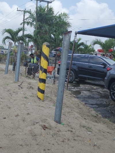 The scene, located in Igando Oloja, Nigeria, depicts an outdoor area characterized by sandy terrain in the foreground sloping downward from left to right. Three vertical poles are present; one prominent yellow and black striped pole stands in the mid-left, flanked by two bare grey metal poles. Small bits of debris and sparse vegetation are visible on the sand. In the midground, a male individual with dark skin and hair, wearing a light shirt under a lime green reflective vest, is observed bending over. He is actively interacting with multiple dark fabric items, possibly garments or bags, hanging on a low structure. To the right, a dark-colored SUV is partially parked on a wet, dark surface, with the rear of another lighter-colored vehicle visible adjacent to it. A blue canopy extends from the right, partially shading the vehicles. The background features dense green palm trees and other foliage, along with overhead electrical poles and wires. Distant structures are faintly discernible behind the vegetation. The sky is partly cloudy.