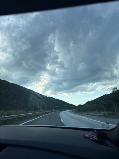 A high-angle, wide shot captures a road winding through a mountainous landscape. The sky is filled with dramatic, dark clouds, but shafts of sunlight break through in places. The road, with a white line marking its edge, curves to the right and disappears into the distance. On either side of the road, steep, tree-covered hills rise up, their slopes darkened by the overcast sky. A metal guardrail runs along the side of the road. A dark dashboard with a small cluster of dried flowers and keys is visible in the foreground. The scene appears to be a rural highway in the vicinity of Dolno Varshilo, Bulgaria.