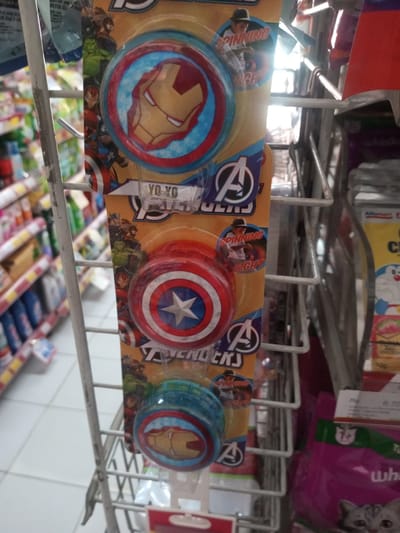 A close-up shot captures a display of Avengers-themed yo-yos hanging on a wire rack in what appears to be a retail setting, possibly a supermarket or convenience store. The yo-yos feature designs of Iron Man and Captain America. The packaging has the Avengers logo on it, as well as a picture of a person using a yo-yo. The background includes shelves stocked with various items, suggesting a store aisle. The location details (Zeyathiri District, Myanmar) are not visually evident in the image.