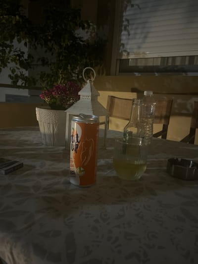 The image depicts a table with various items on it, set outdoors in what appears to be a patio or balcony area. The lighting suggests it is evening or nighttime. In the foreground, there is a can of what looks like orange-flavored soda, a glass with a yellow liquid, a bottle of water, and an ashtray. To the left, a pack of cigarettes is visible. In the background, there is a potted plant with purple flowers, a white decorative lantern, and chairs. A building with blinds is also visible in the background. The overall scene is consistent with a relaxed setting, possibly a bar or restaurant in a coastal area. The location is identified as Ofrynio Beach, Greece.
